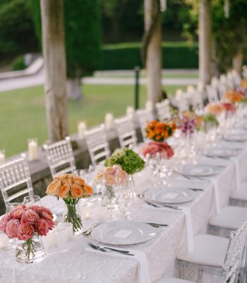 A very long table for guests with a white tablecloth, floral arrangements, glass plastic transparent chairs Chiavari. Under the old columns with vines of wisteria.